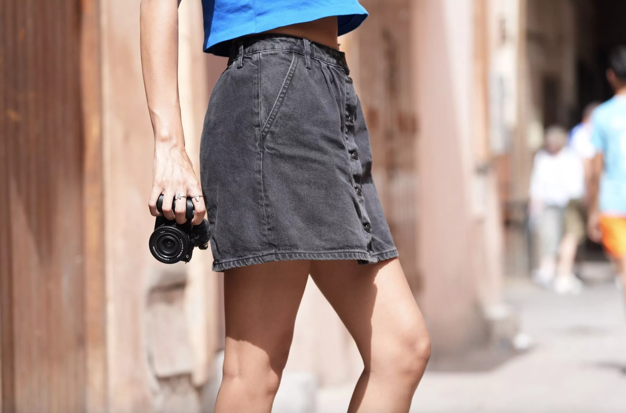Kobieta trzymająca czarny aparat, ubrana w niebieski top i szarą spódnicę, na ulicy.