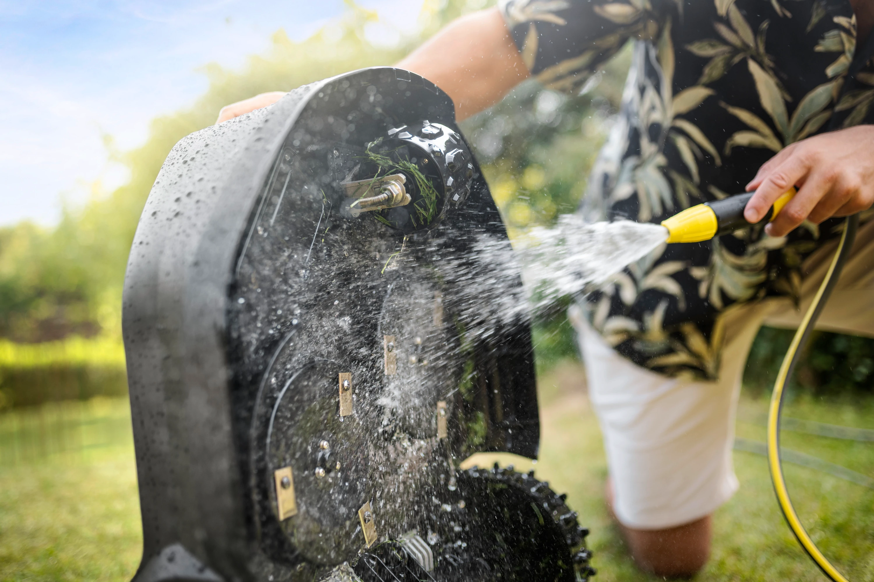 Osoba czyści kosiarkę wodą. Kosiarka jest czarna. Niebo jest niebieskie, trawa jest zielona.