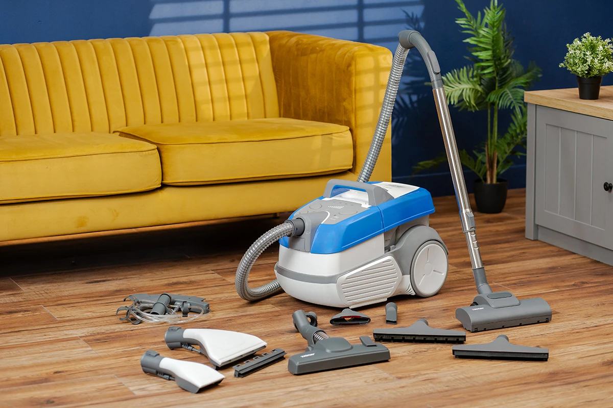 Odkurzacz z akcesoriami na drewnianej podłodze, żółta sofa, roślina i szara szafka w tle.
