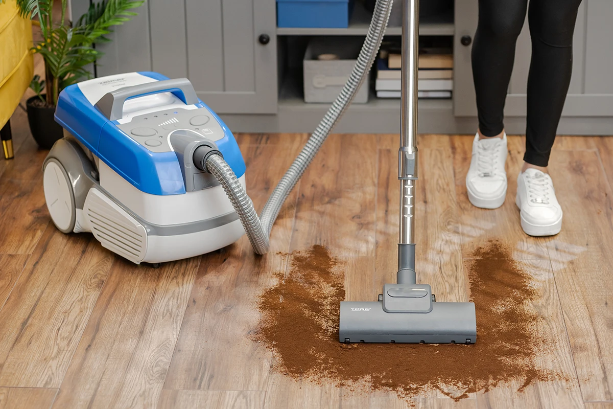 Odkurzacz na drewnianej podłodze, czyszczenie brązowych zanieczyszczeń, osoba stoi.