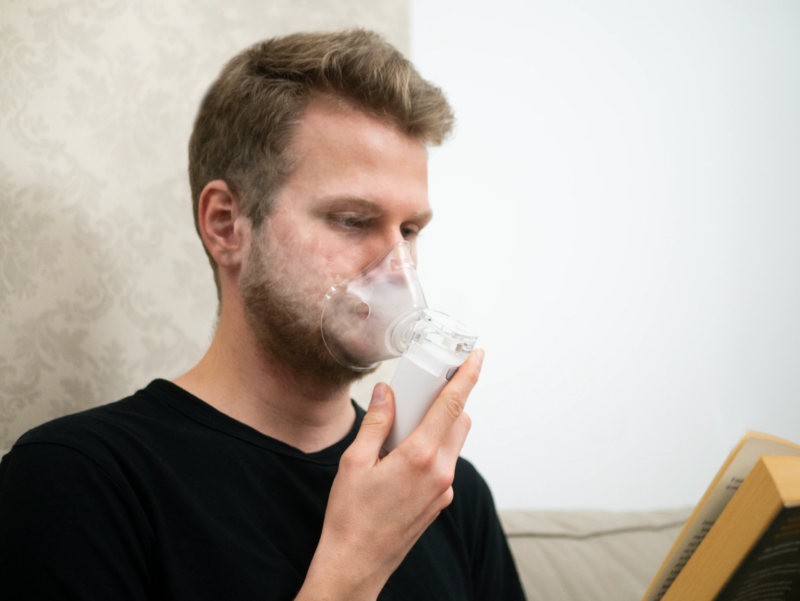 Mężczyzna w masce nebulizatora czyta książkę. Trzyma urządzenie. Tłem jest ściana i kanapa.