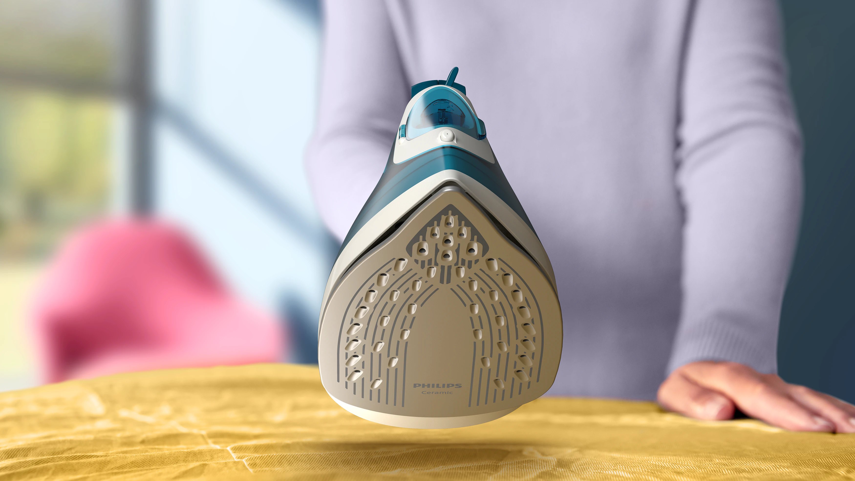 Niebiesko-białe żelazko Philips na żółtej tkaninie. Widoczna dłoń i ramię osoby. Tło jest rozmyte.