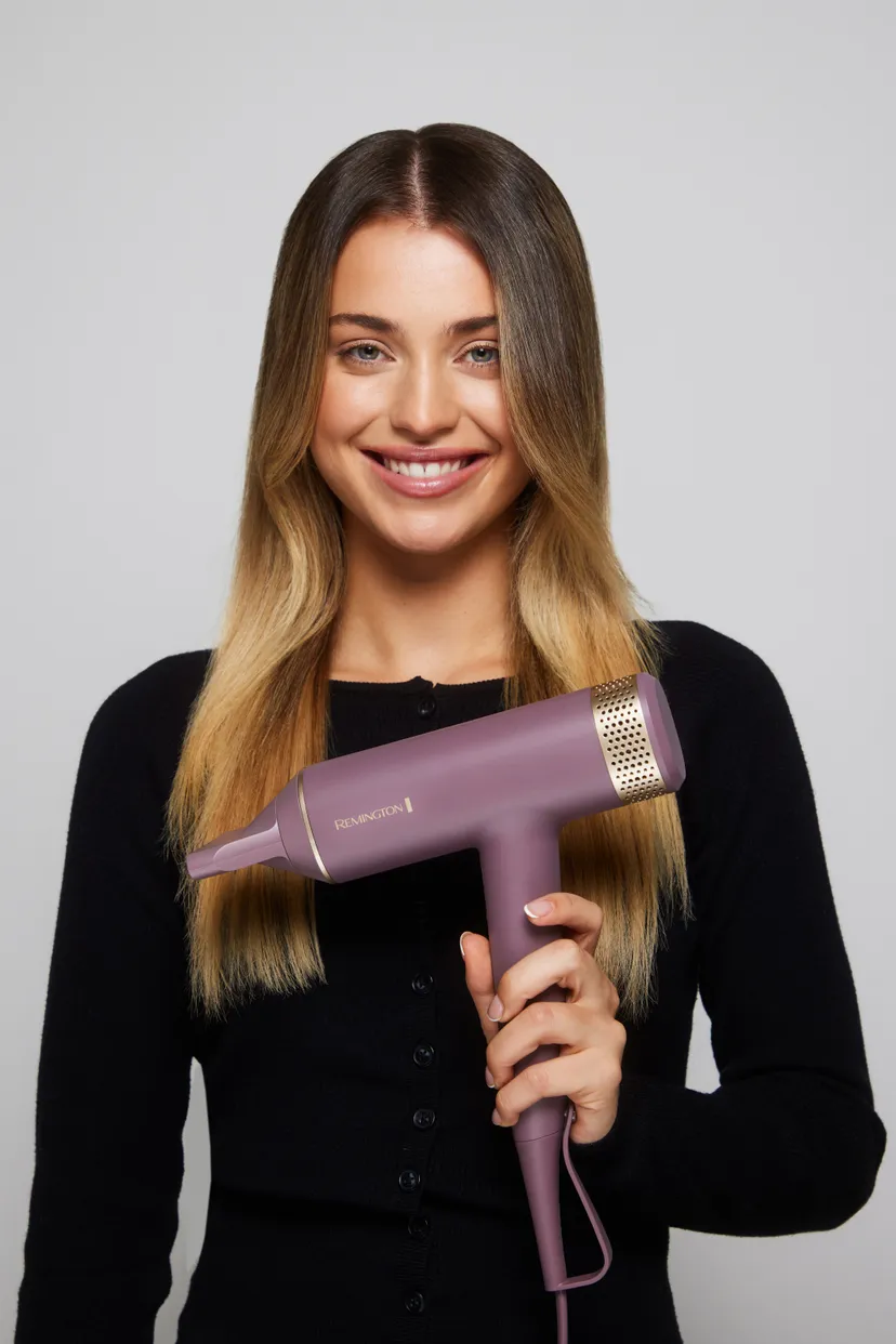 Kobieta trzyma fioletową suszarkę do włosów. Uśmiecha się, z długimi blond włosami, w czarnym swetrze, na szarym tle.