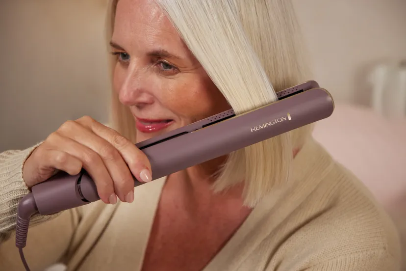 Kobieta z białymi włosami używa prostownicy Remington. Prostownica jest fioletowa. Jest w pokoju z różowym tłem.