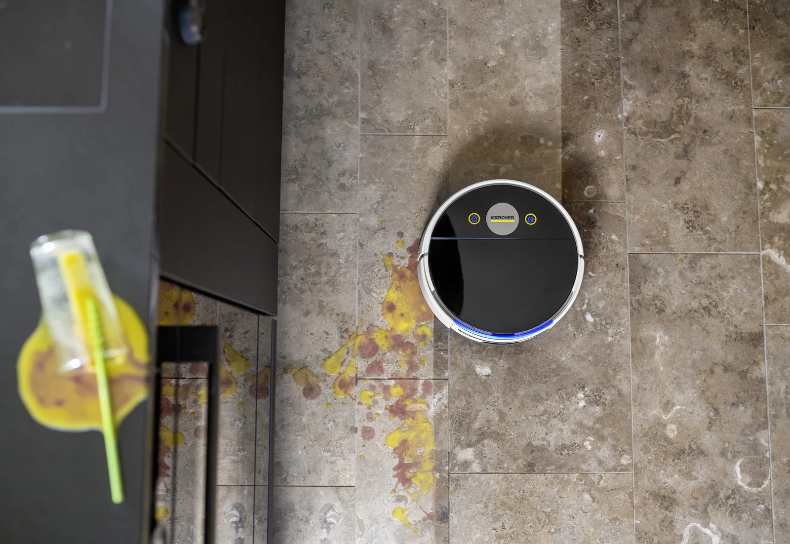 Rozlana woda na podłodze z płytek, obok odkurzacz robot. Obecna jest również szklanka ze słomką.