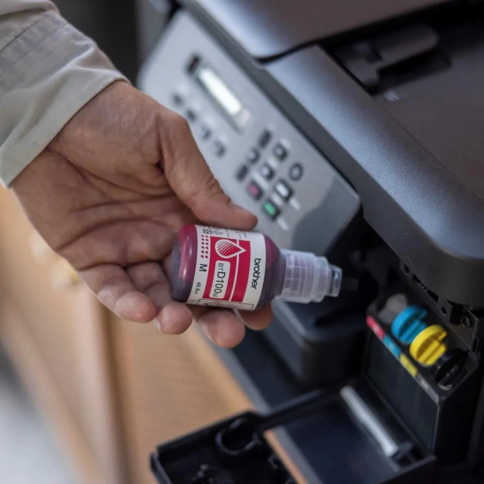 Ręka trzyma butelkę z magenta tuszem nad otwartym gniazdem na wkład drukarki. Drukarka jest czarna, a butelka ma białą etykietę.