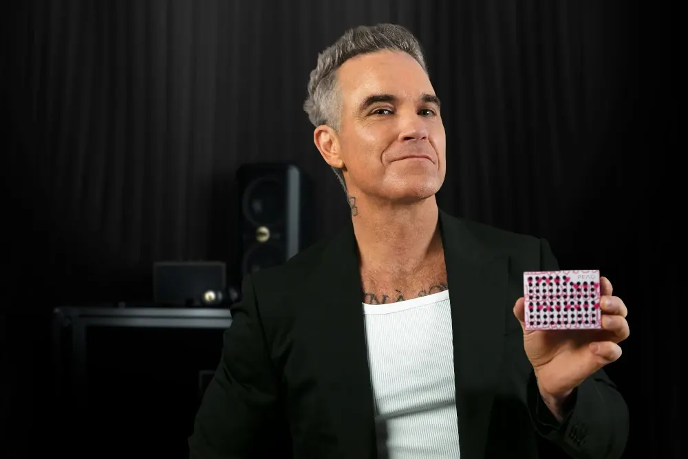 Mężczyzna z siwymi włosami w czarnym garniturze trzyma małe białe pudełko z różowymi kropkami.