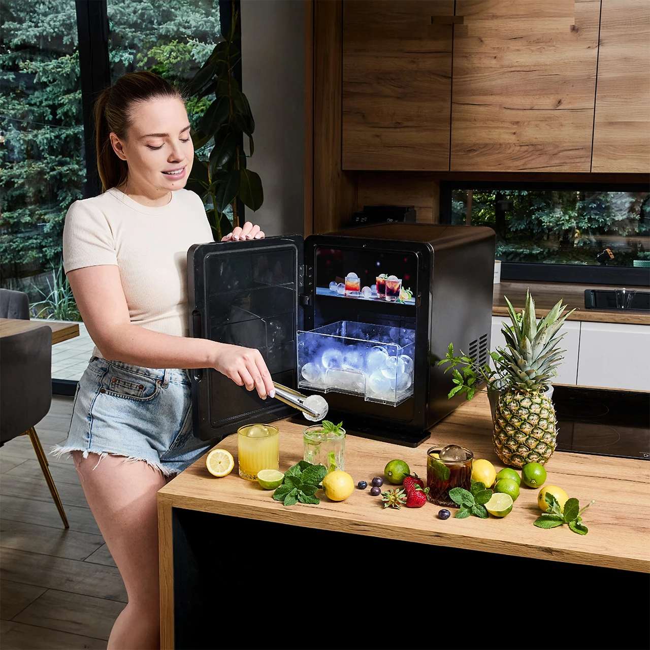 Kobieta z szczypcami do lodu otwiera mini lodówkę. Napoje, owoce i ananas na blacie. Tłem jest kuchnia z drewnianymi szafkami.
