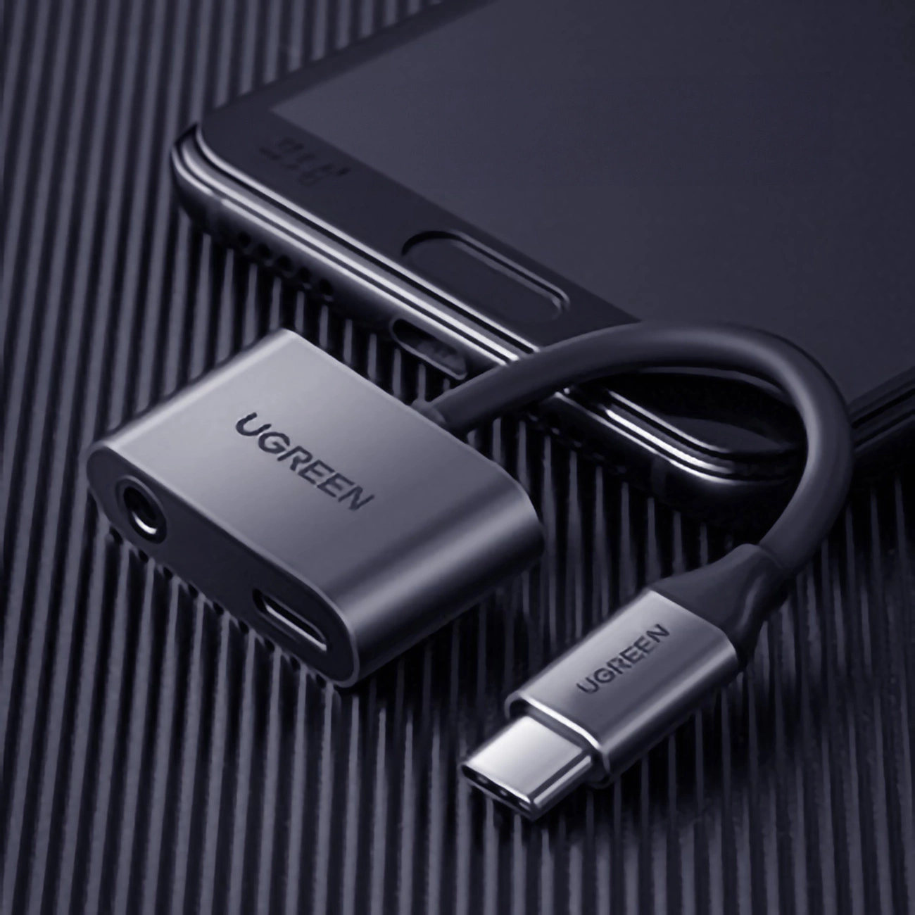 Adapter UGREEN z złączem USB-C i gniazdem słuchawkowym, podłączony do smartfona. Adapter i telefon są ciemnoszare.