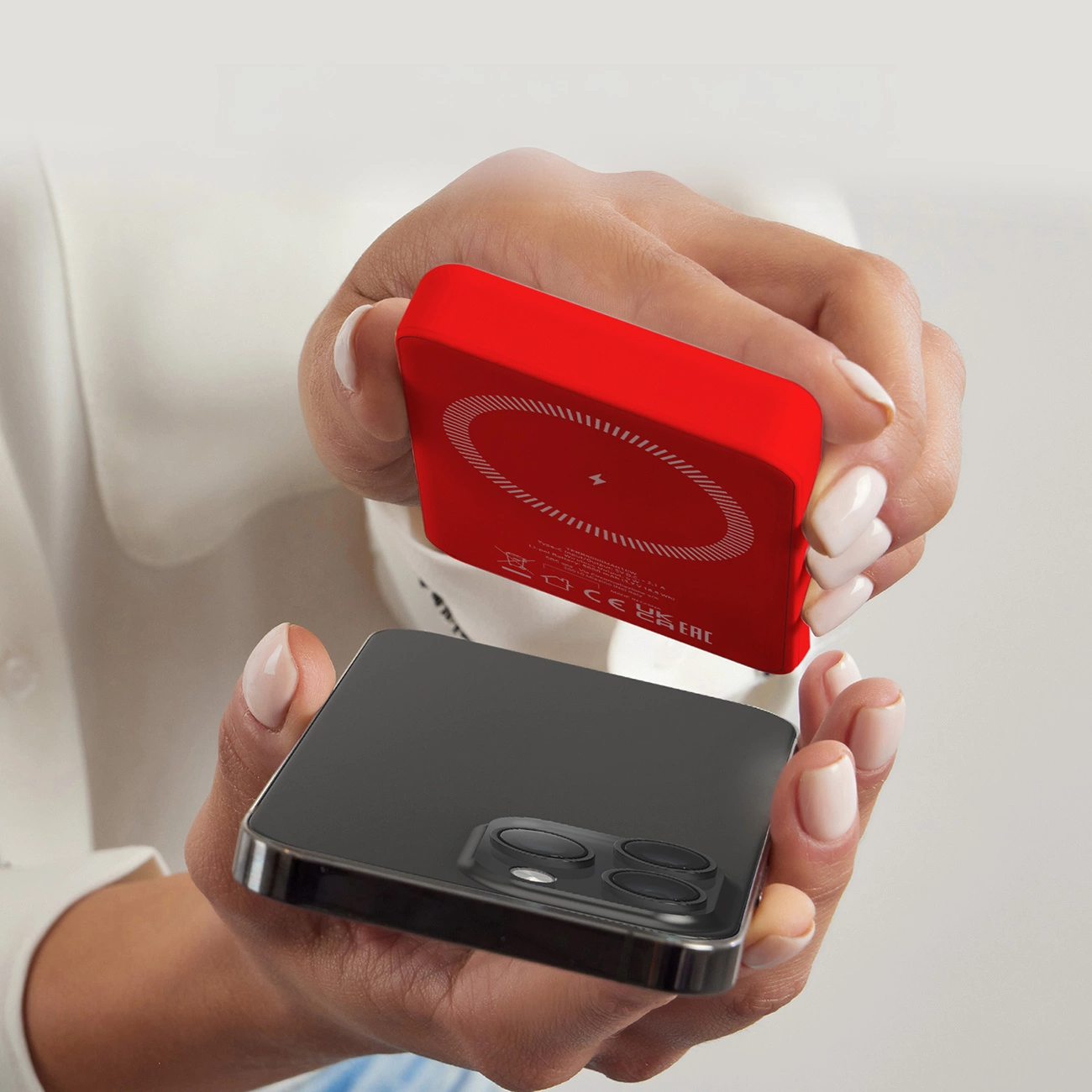 Dłonie trzymają czarny smartfon i czerwoną ładowarkę bezprzewodową. Ładowarka jest nad telefonem.