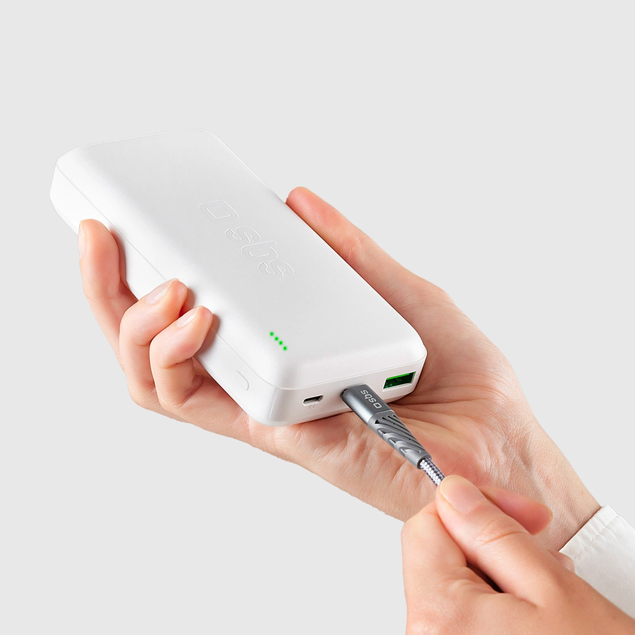 Ręka trzyma biały power bank z wpiętym kablem. Power bank ma logo i zielone wskaźniki.