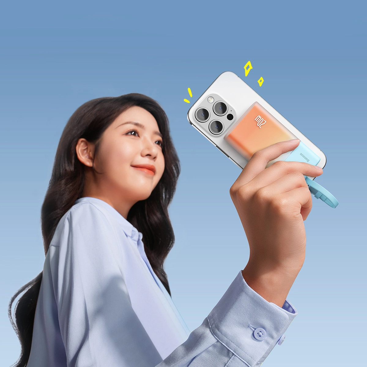 Kobieta trzyma telefon z ładowarką. Telefon jest biały, ładowarka pomarańczowo-niebieska. Uśmiecha się na tle błękitnego nieba.