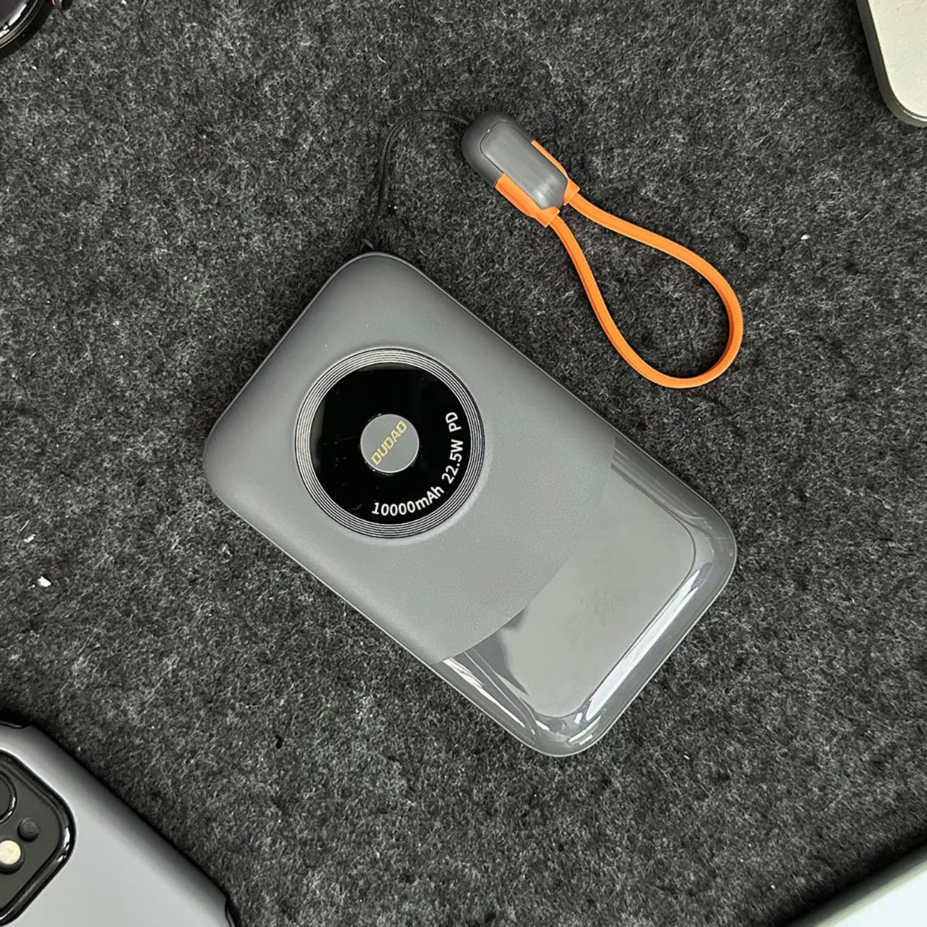 Szary power bank z pomarańczową pętlą leży na szarej powierzchni. Ma czarne koło z białym tekstem: 10000mAh 22.5W PD.