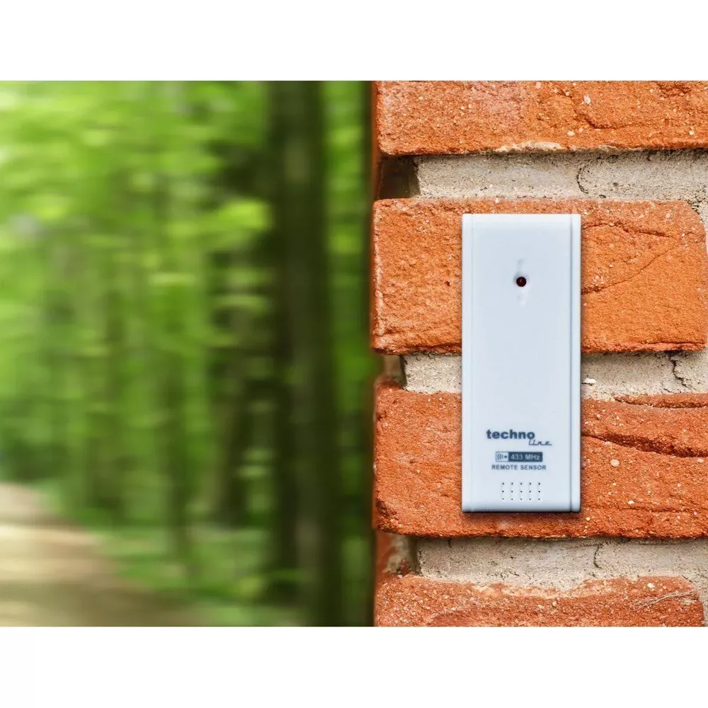 Biały czujnik zdalny z czerwoną kropką przymocowany do ceglanej ściany. Zielone drzewa w tle.