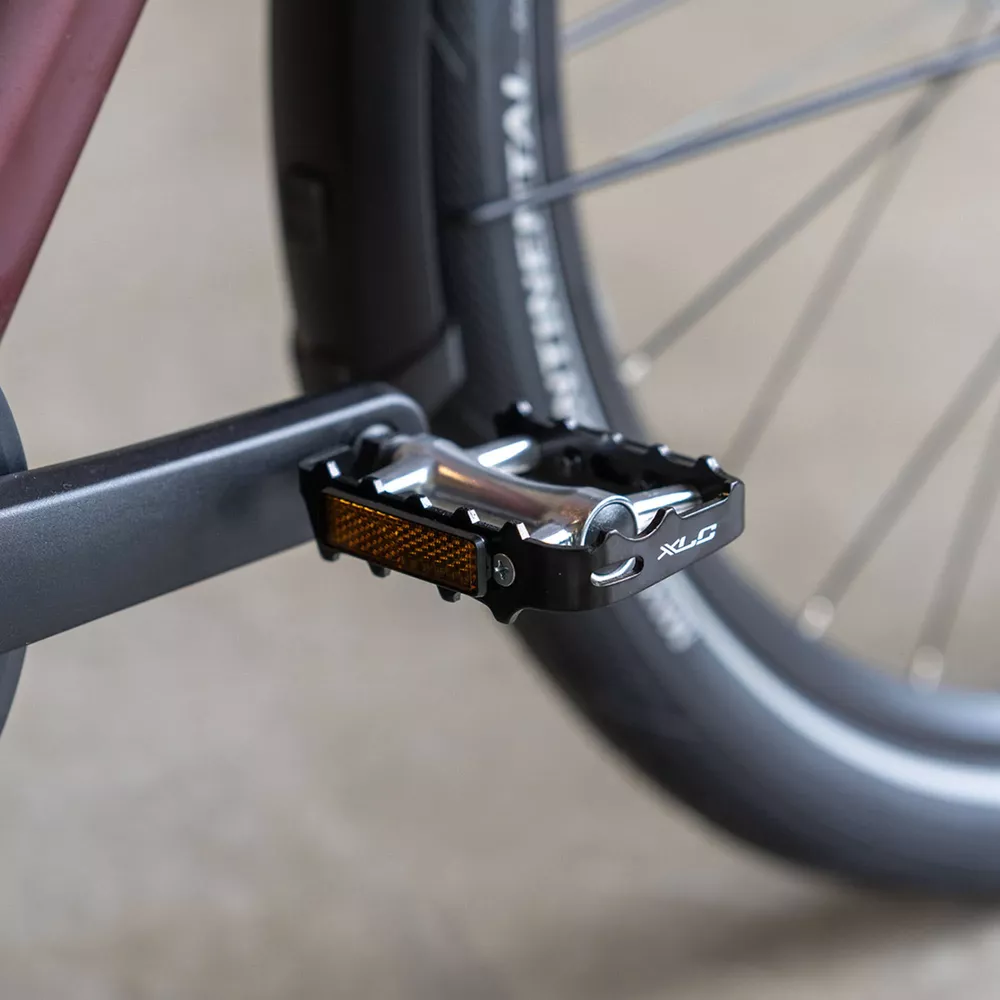 Czarny pedał rowerowy z odblaskiem i logo XLC, obok koła.