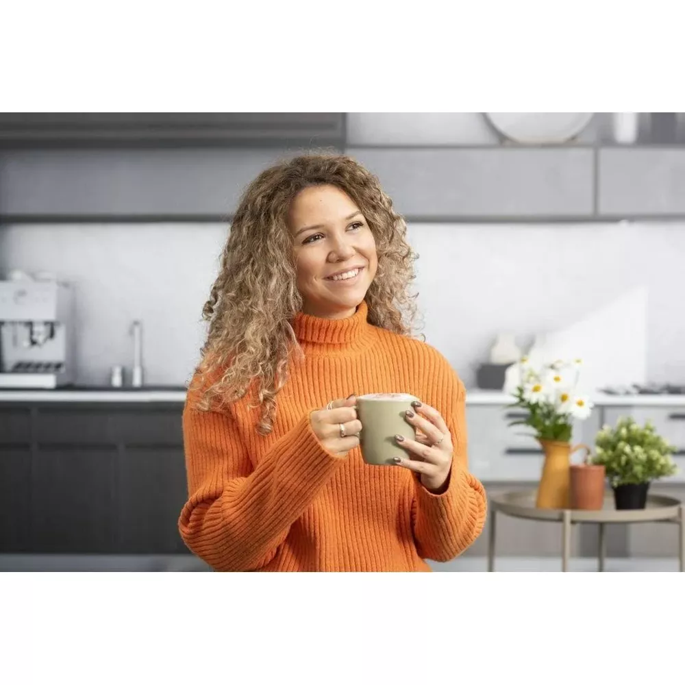 Kobieta z kręconymi włosami w pomarańczowym swetrze trzyma kubek w kuchni.