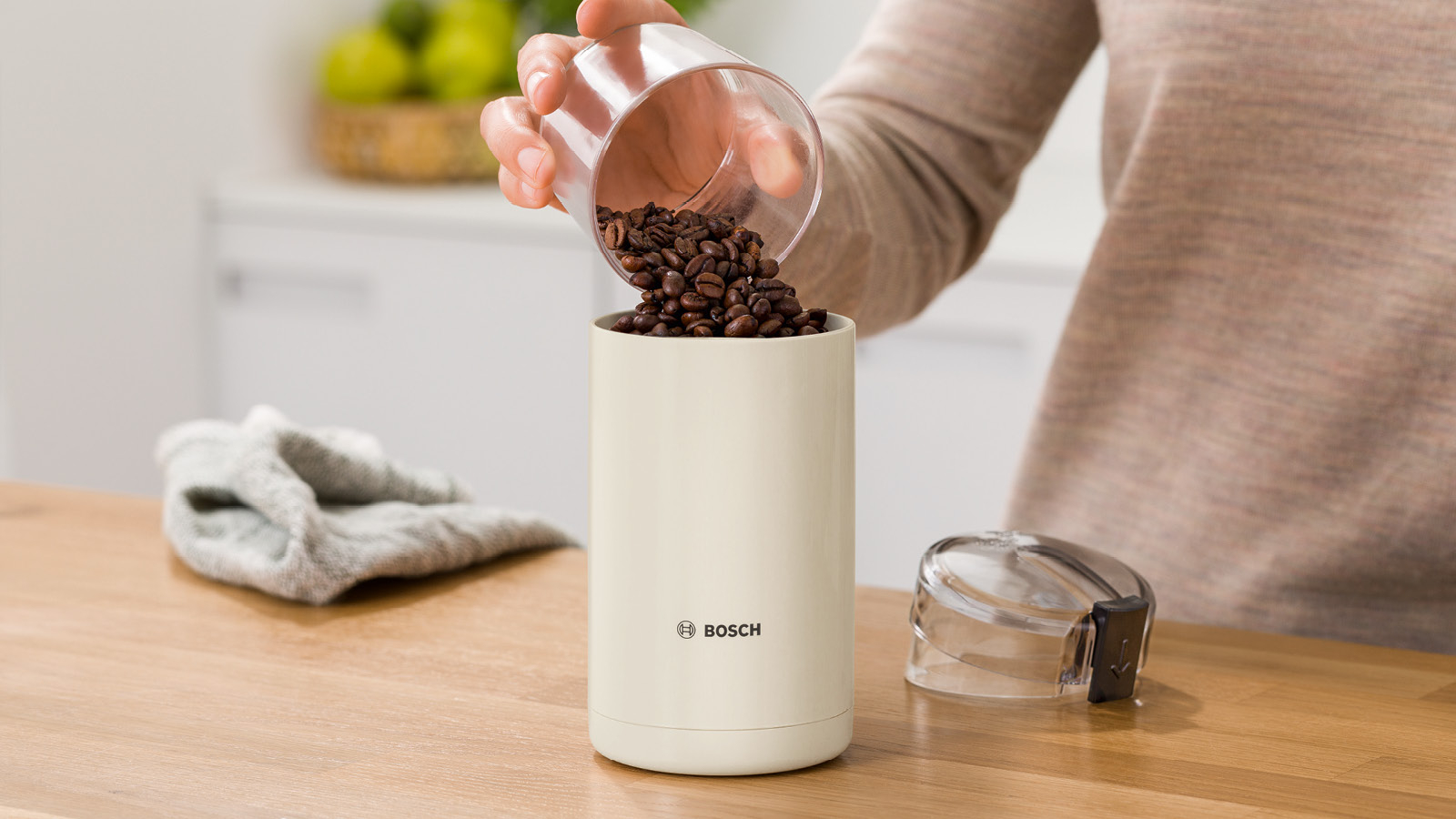 Eine Person schüttet Kaffeebohnen in eine cremefarbene Mühle. Ein Tuch und ein Deckel sind auch da.