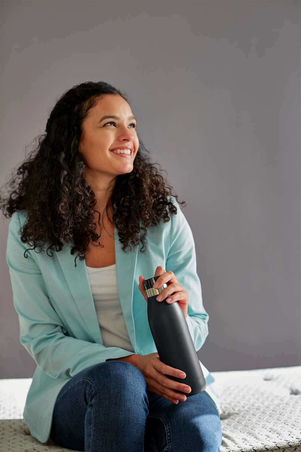 A smiling woman holding a black water bottle, looking up, sitting on a bed.