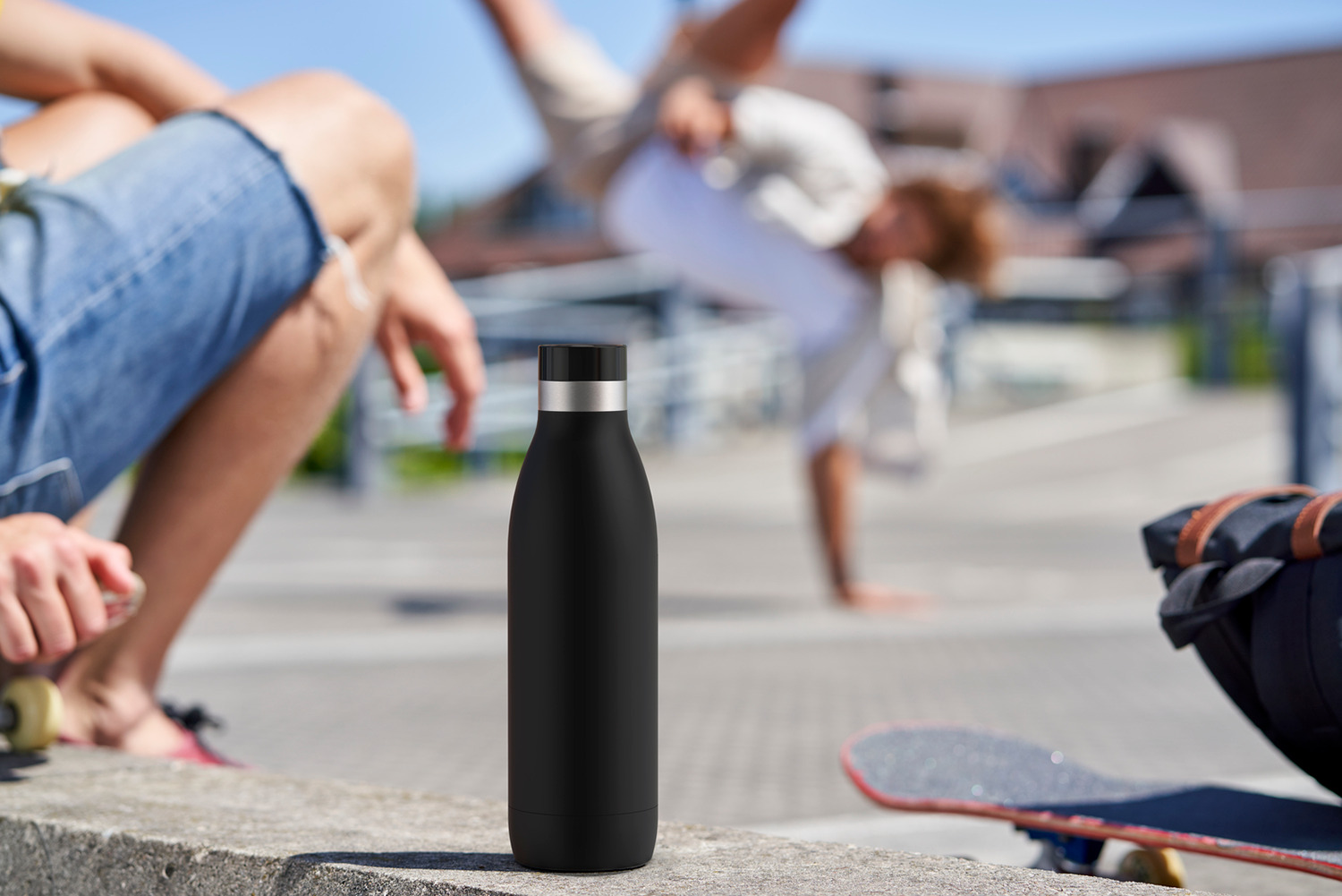 A black water bottle stands on a wall with two people, one skateboarding, in the background.