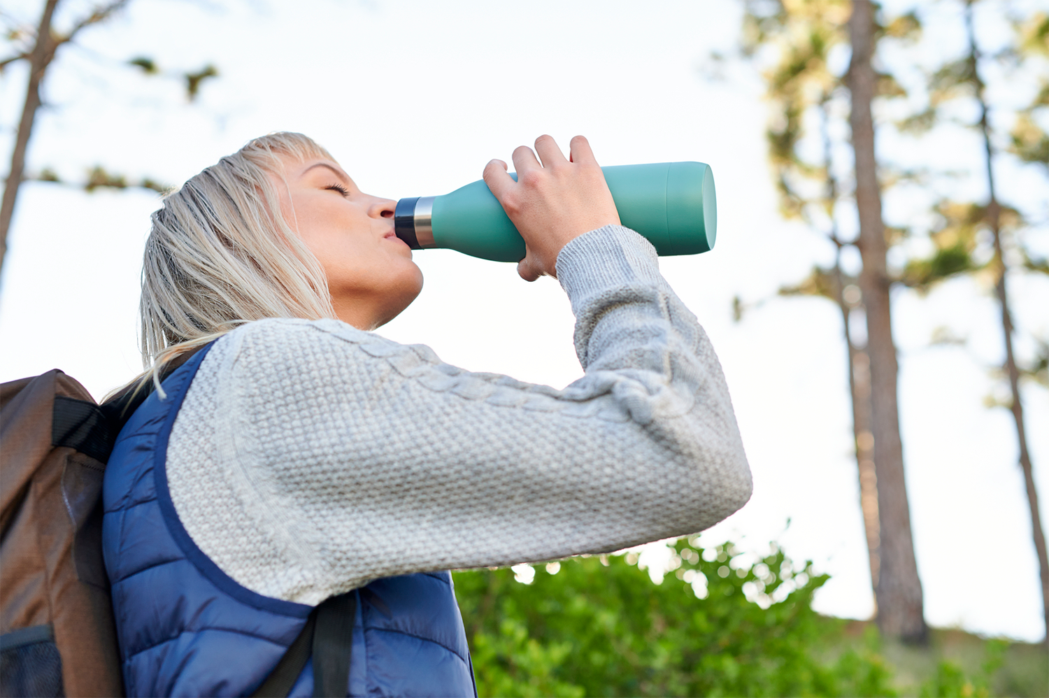 Een vrouw drinkt uit een turquoise waterfles in een bos. De achtergrond is wazig.