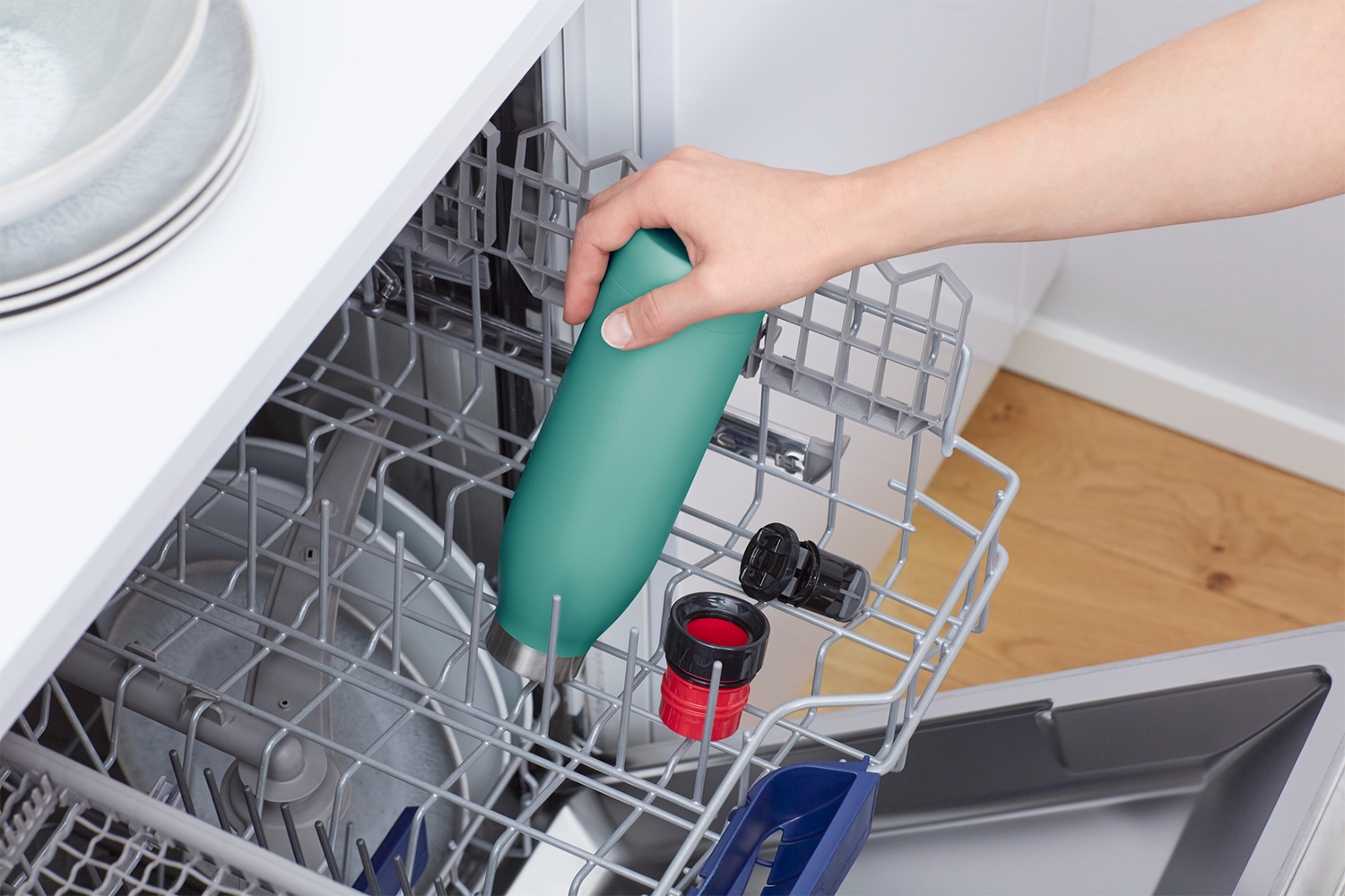 Een vrouw die een turquoise waterfles in een vaatwasser mand zet. Andere items zitten ook in de mand.