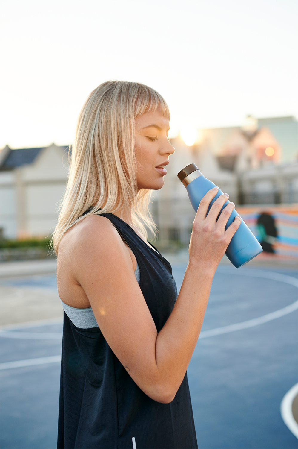 Blonde woman drinking from a blue water bottle, standing on a basketball court.