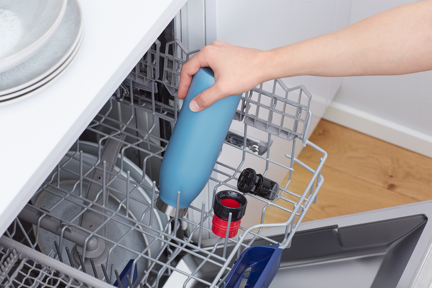 A hand places a blue water bottle in a dishwasher rack with other items.
