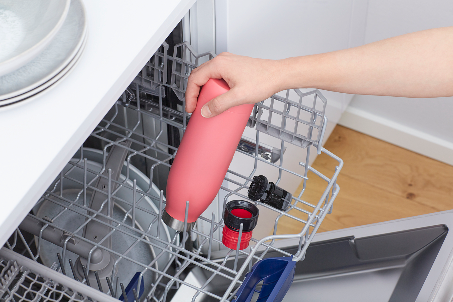 A hand placing a pink water bottle into a dishwasher rack with other items.