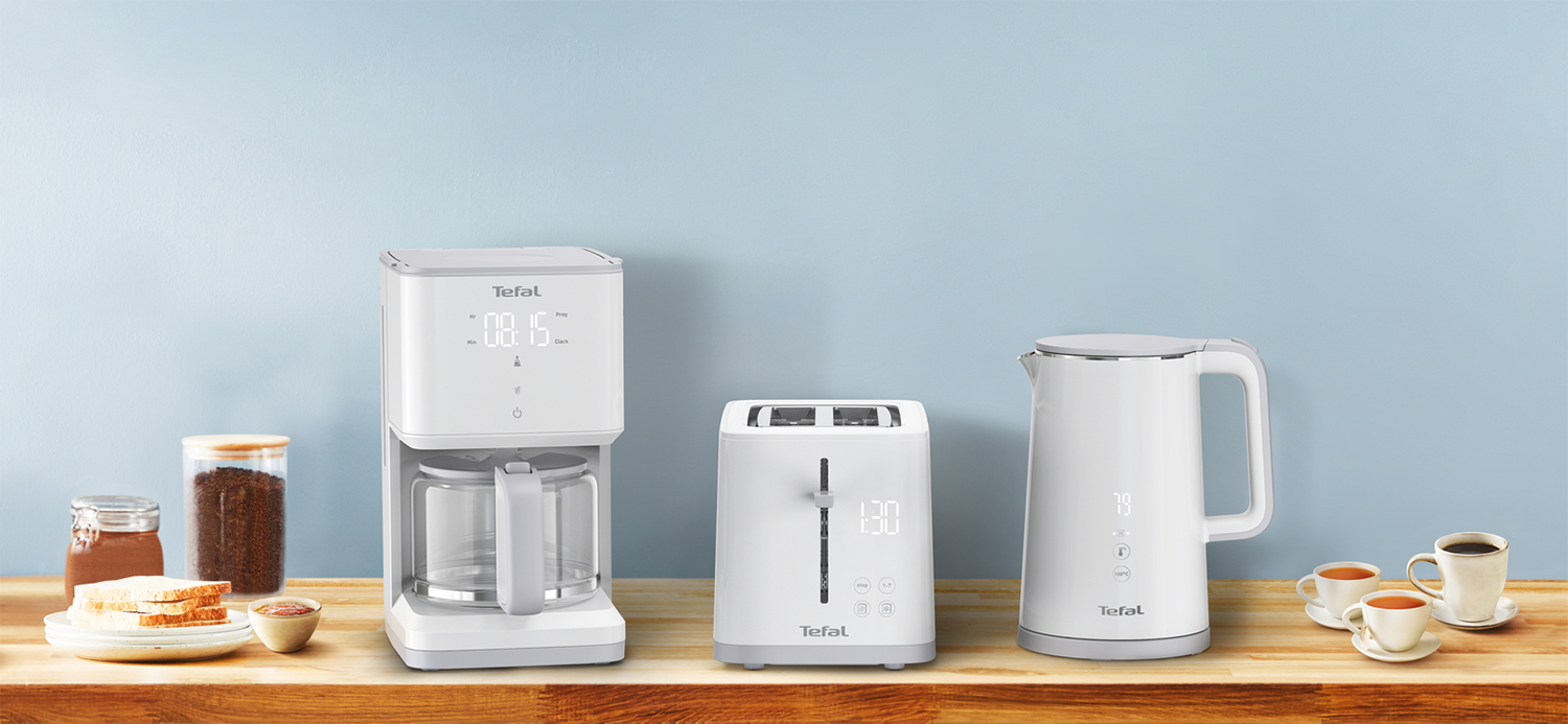 White kitchen appliances on a wooden counter. Coffee maker, toaster and kettle. Cups and ingredients are present.