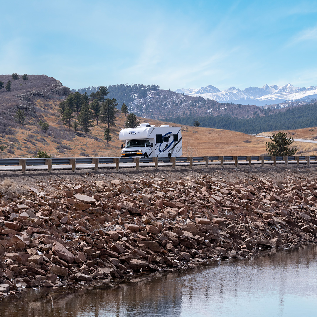 Un camping-car blanc roulant sur une route à côté d'un plan d'eau avec des montagnes au loin.