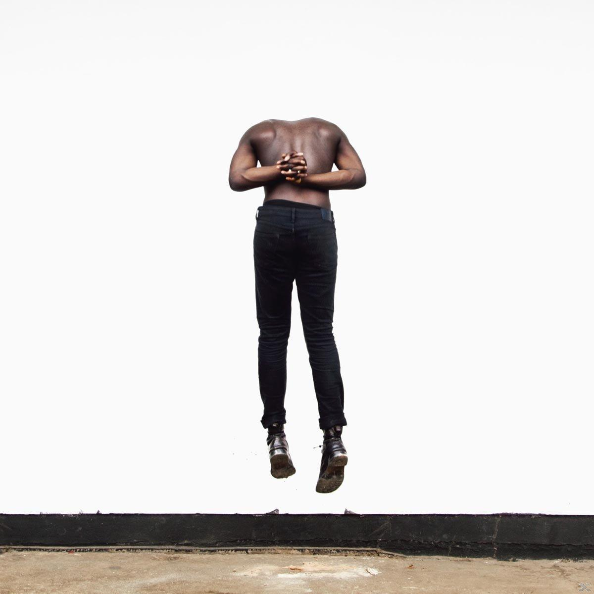Ein kopfloser Mann in der Luft. Arme hinter dem Rücken verschränkt. Trägt schwarze Jeans und Stiefel. Weißer Hintergrund.