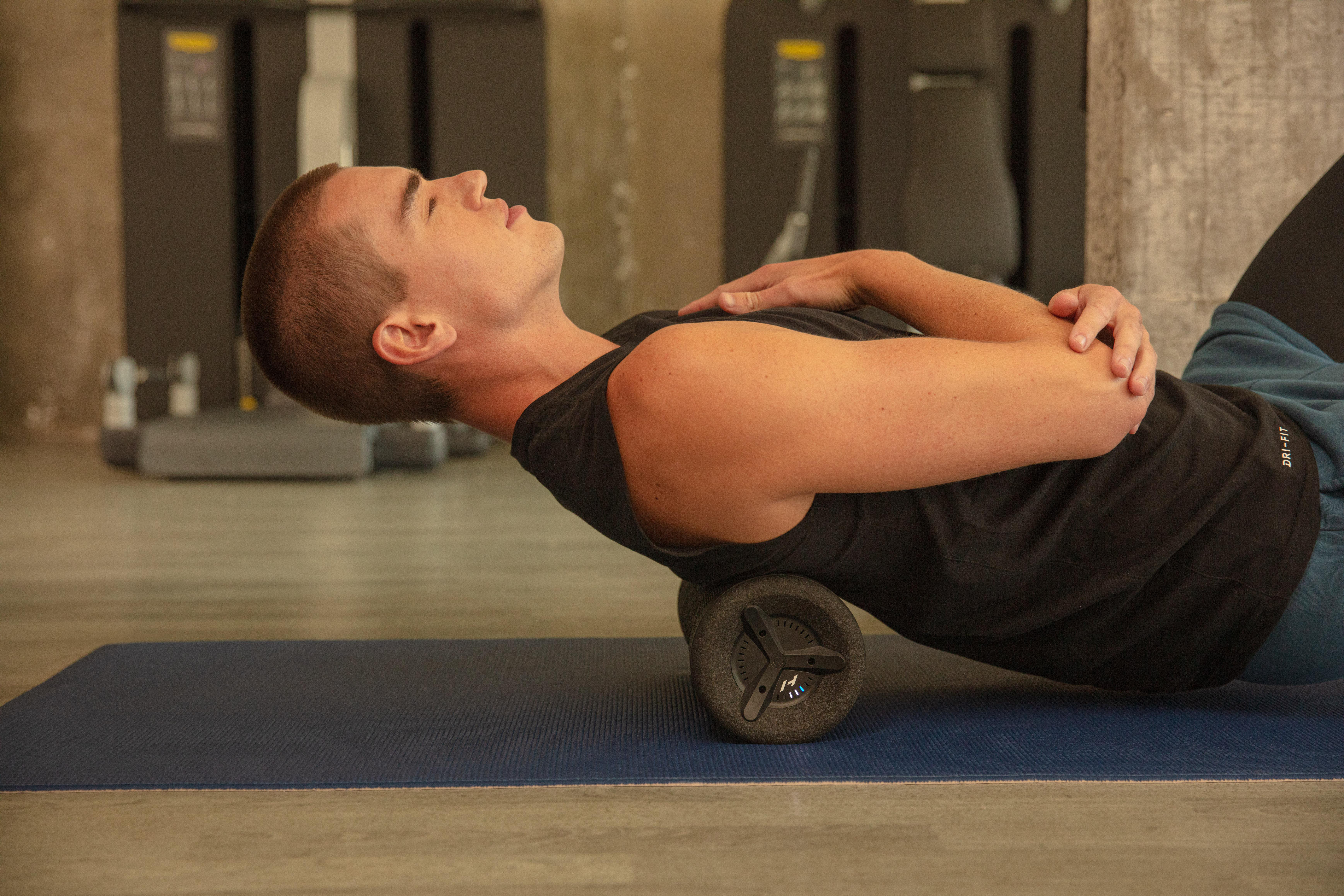 Mann liegt auf einer Trainingsrolle, Rücken gewölbt. Fitnessstudio-Umgebung. Trainingsgeräte vorhanden.