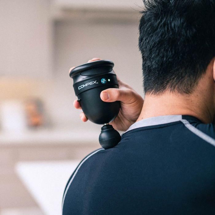 Person verwendet eine schwarze Massagepistole am Rücken einer anderen Person. Compex-Logo sichtbar.