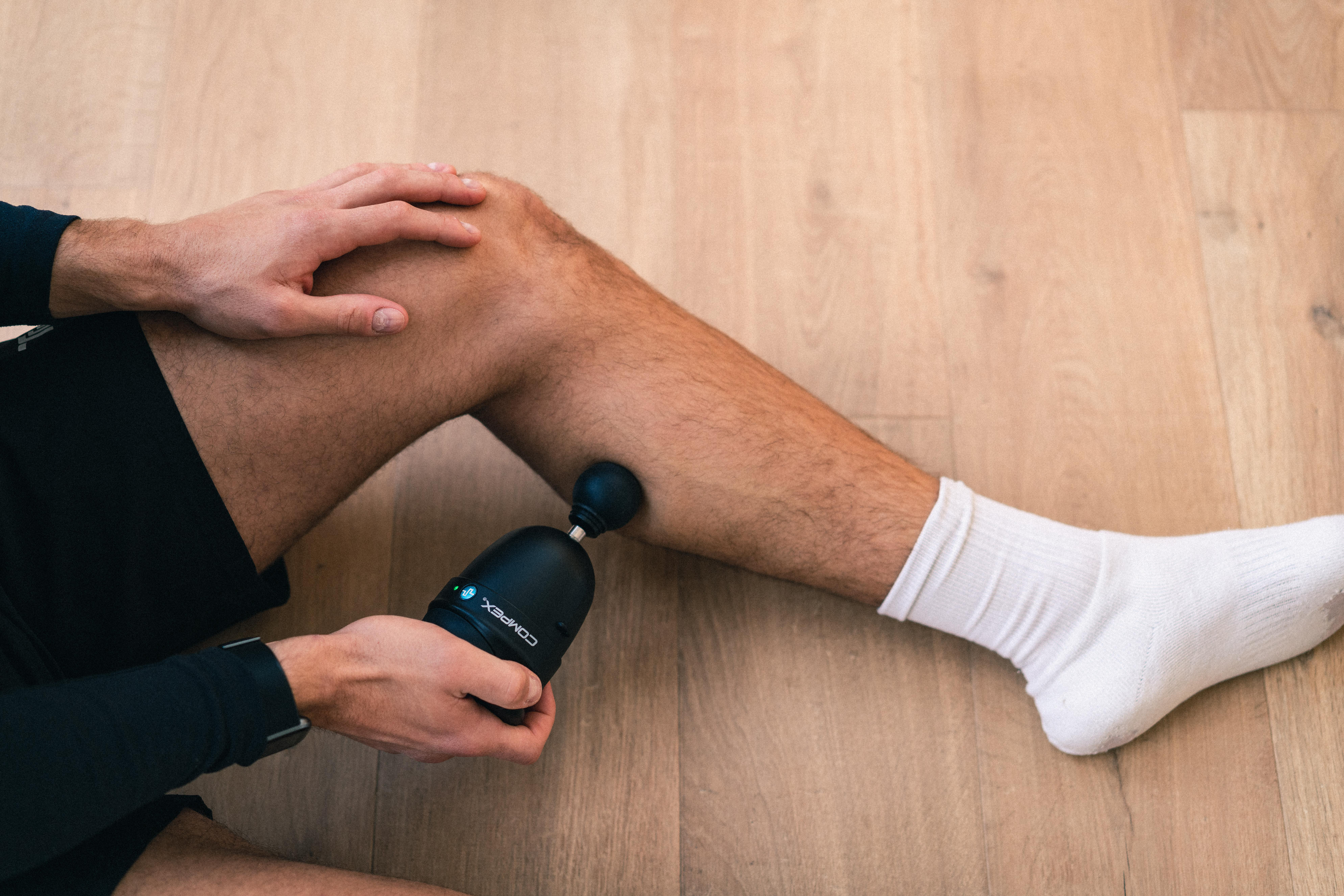 Person verwendet schwarzen Compex-Massagegerät am Bein. Holzfußboden. Weißer Socken.