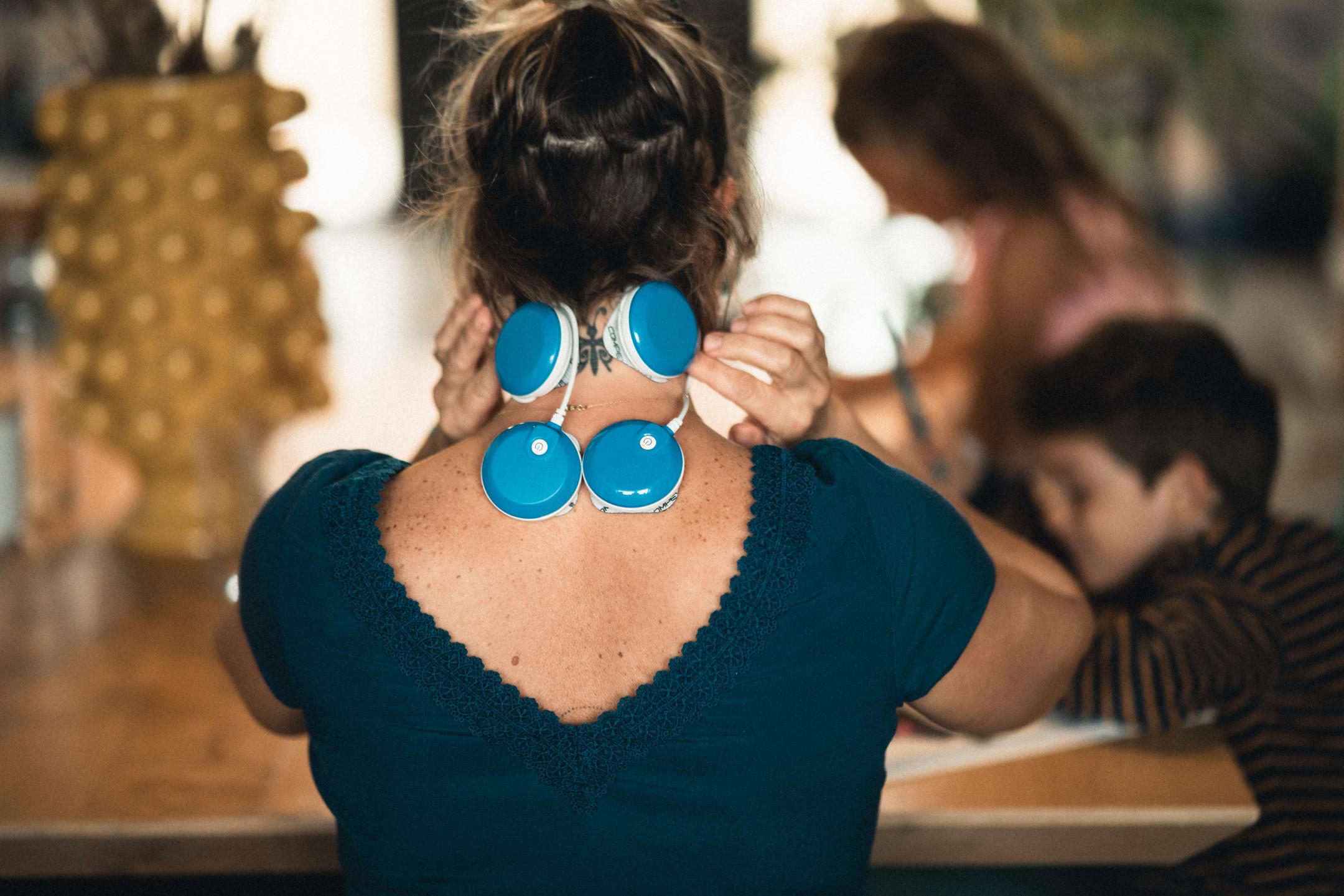 Eine Frau mit einem Massagegerät am Hals, Kinder im Hintergrund verschwommen.