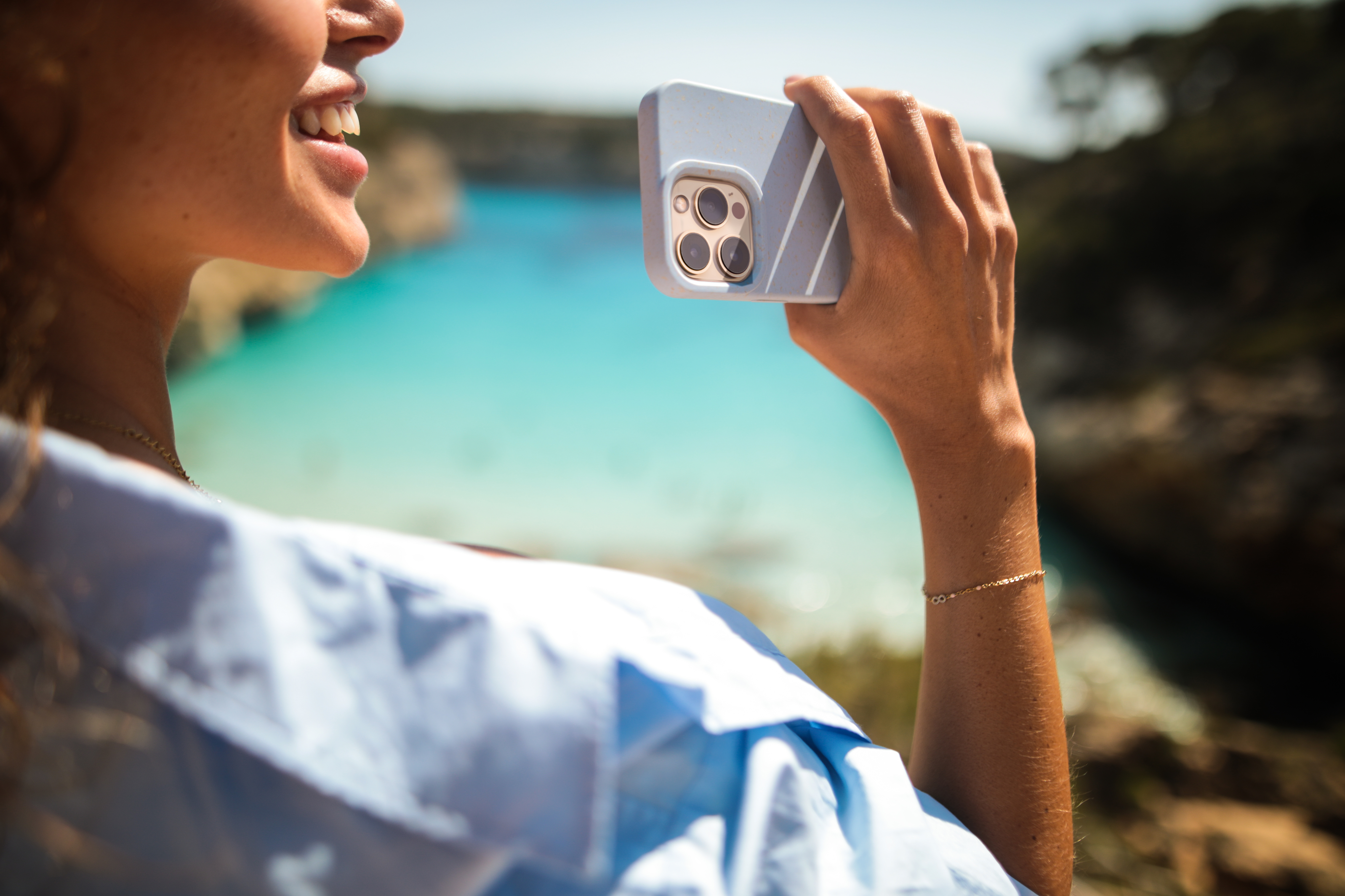Frau hält Telefon und macht ein Foto. Im Hintergrund sind blaues Meer und Felsen zu sehen. Sie lächelt.