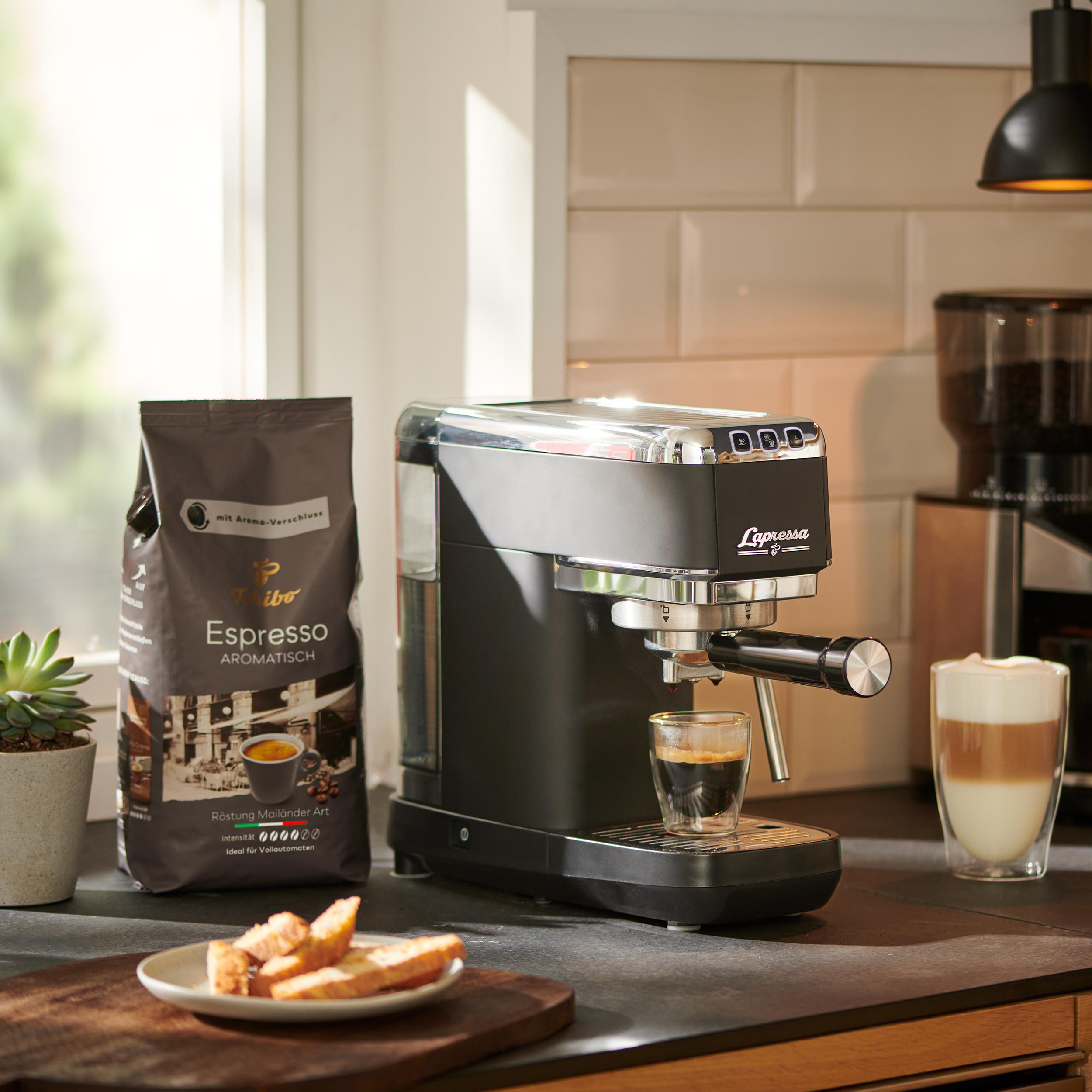 Espressomaschine, Kaffeebohnen und ein Latte auf einer Theke mit Fenster und einer kleinen Pflanze.