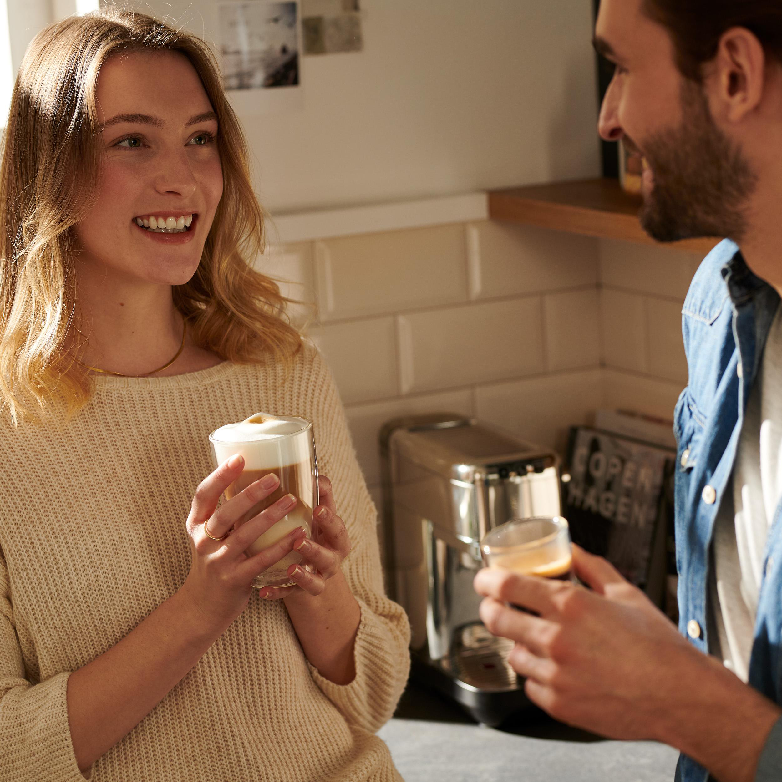 Zwei Personen, eine hält einen Latte, die andere Kaffee. Hintergrund: Küche.