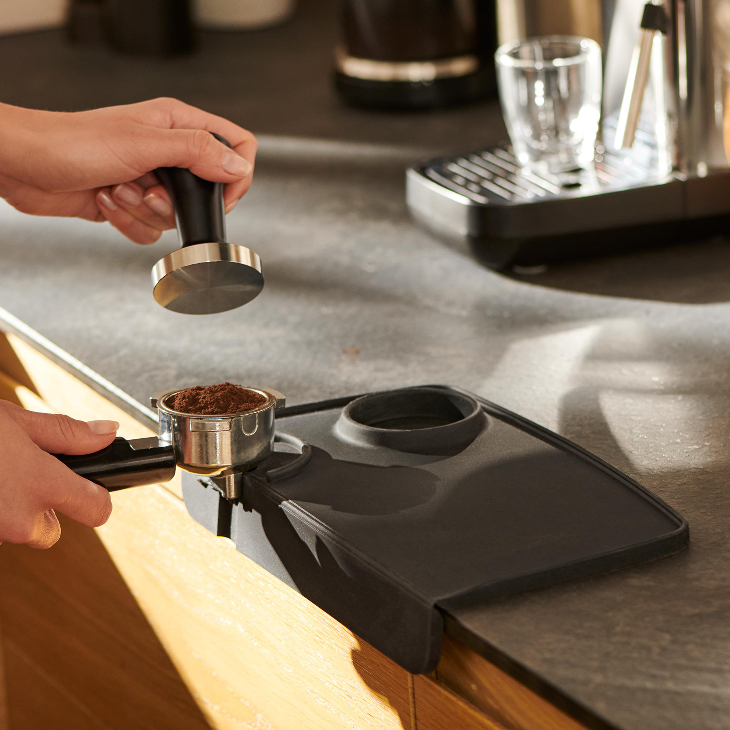 Hände bereiten Espresso zu, Tamper und Pulver, schwarze Matte auf Theke.