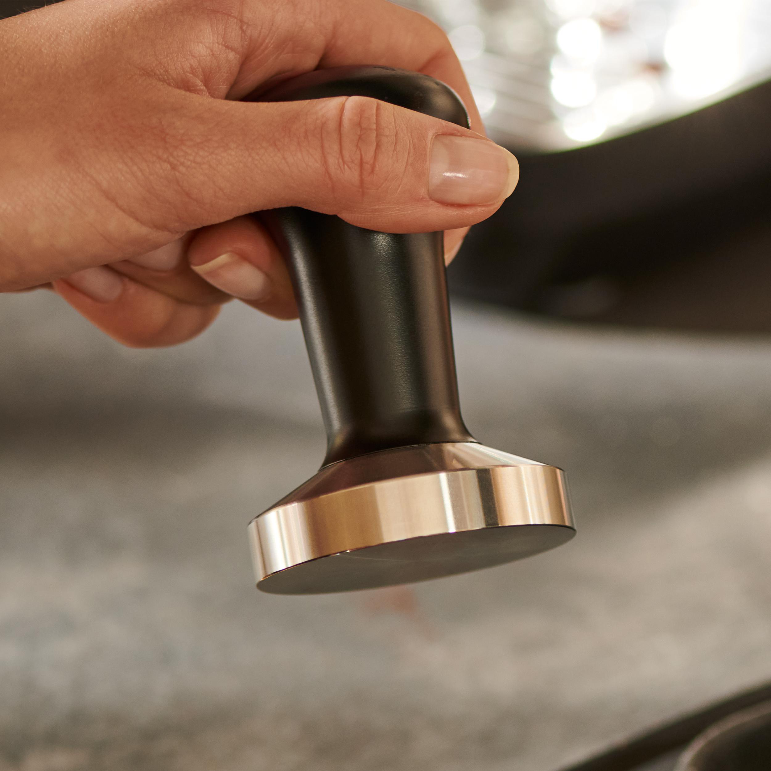 Hand hält einen schwarzen Kaffeestampfer. Silberbasis, Holzgriff. Verschwommener Hintergrund.