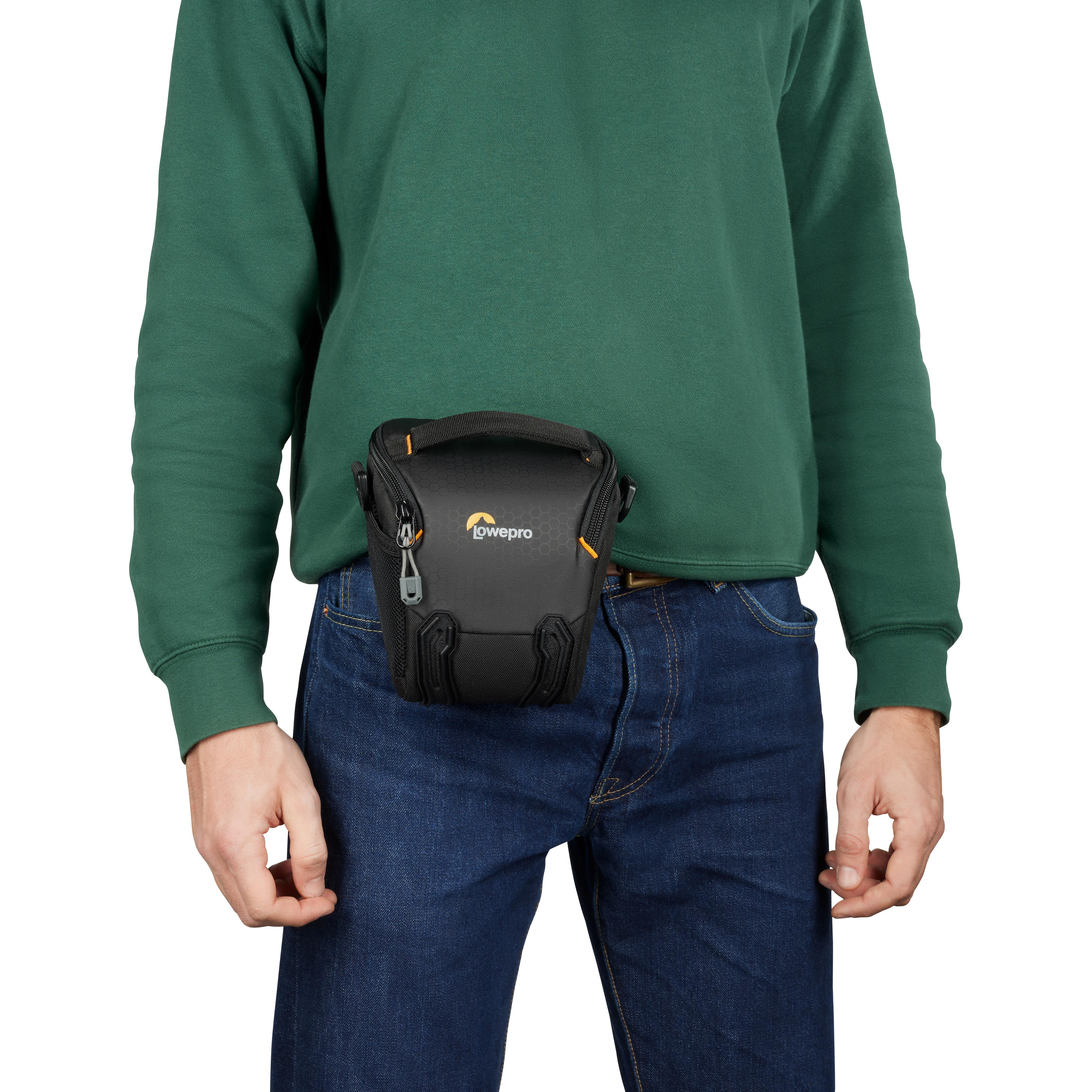 Ein Mann in einem grünen Pullover und einer blauen Jeans trägt eine schwarze Lowepro Kameratasche.
