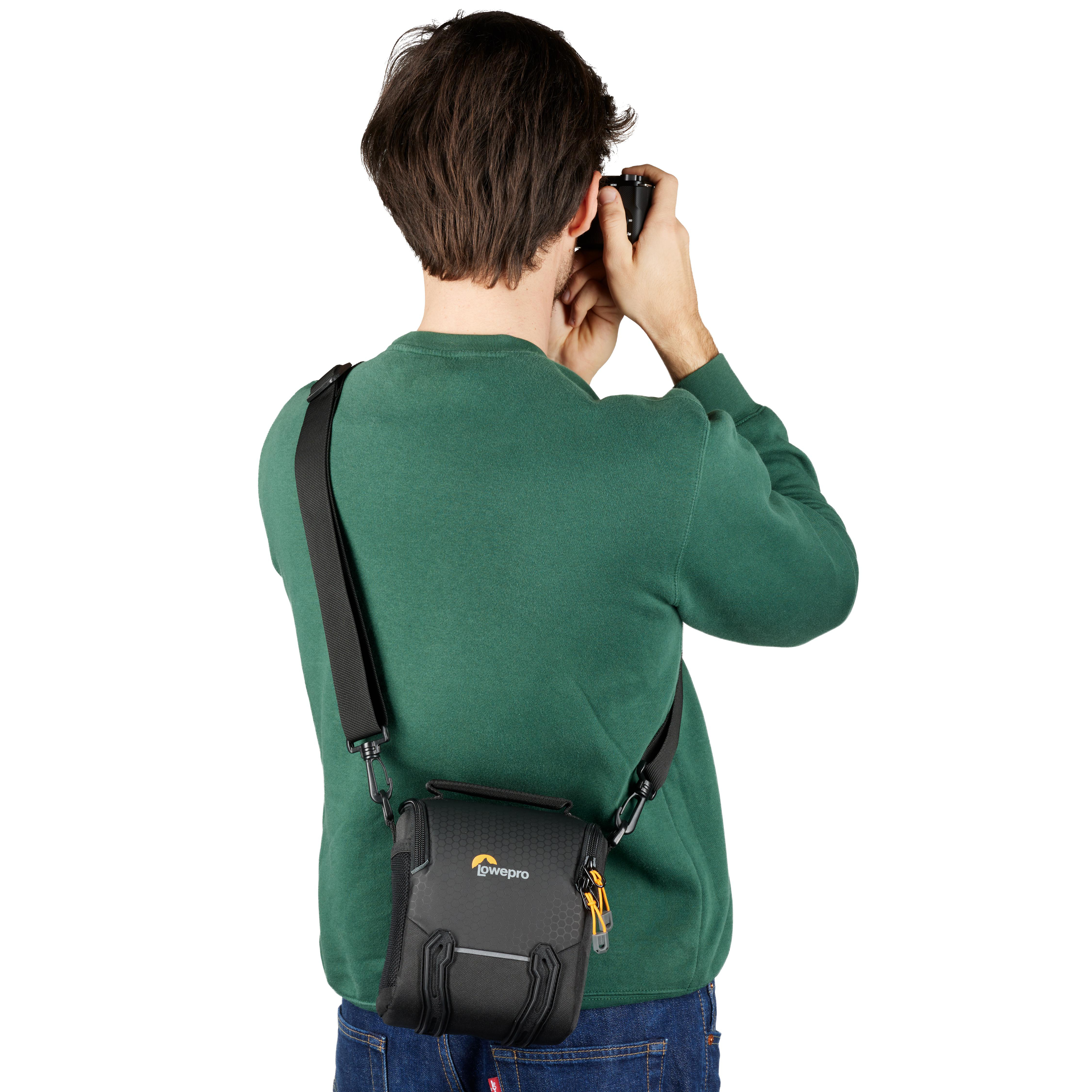 Ein Mann mit grünem Pullover macht ein Foto mit einer Kamera. Er trägt eine Lowepro-Tasche.