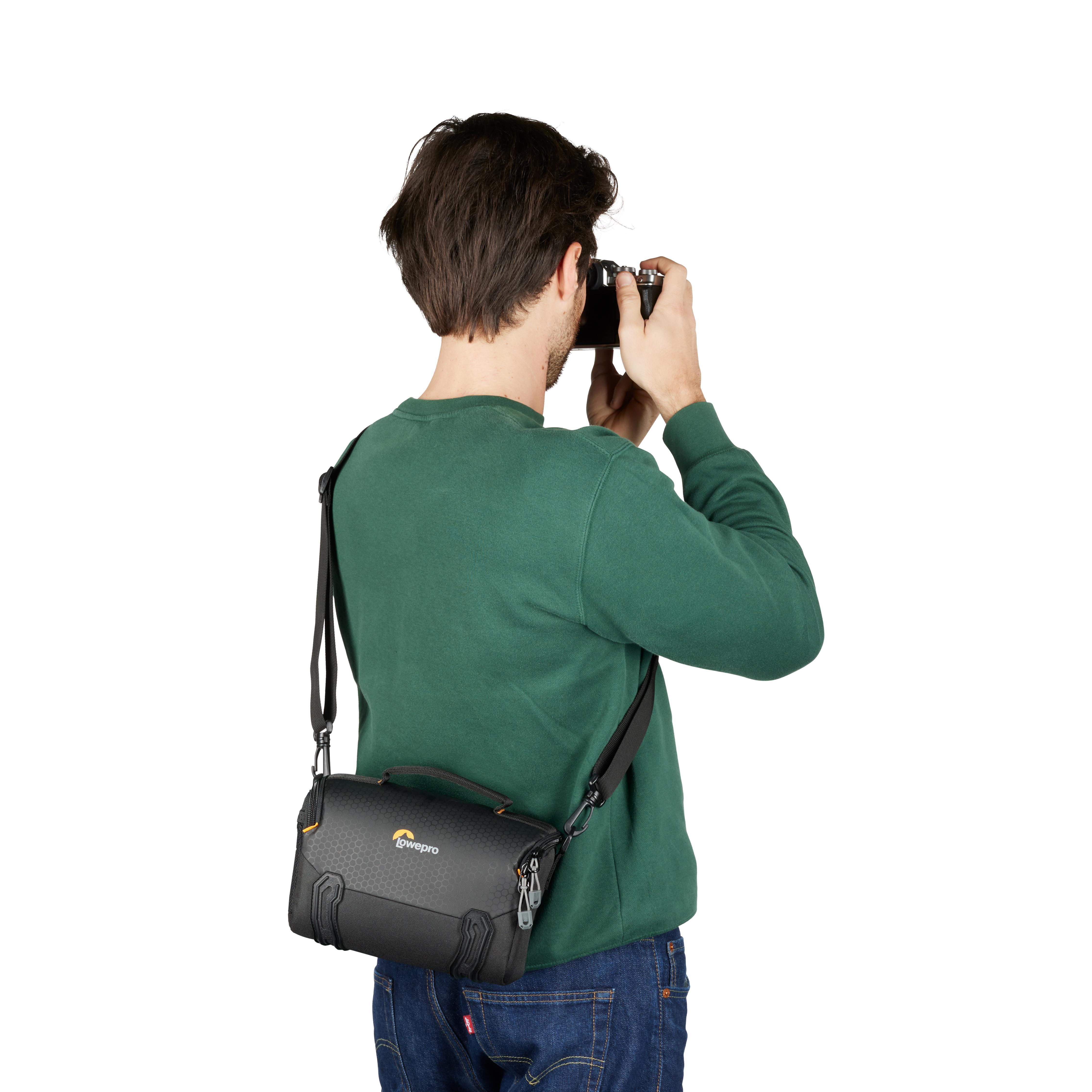 Ein Mann macht ein Foto mit einer Kamera, trägt einen grünen Pullover und eine schwarze Lowepro-Tasche. Weißer Hintergrund.