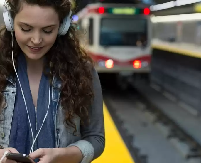 Donna con le cuffie su una banchina, che guarda il suo telefono. Un treno è sullo sfondo.