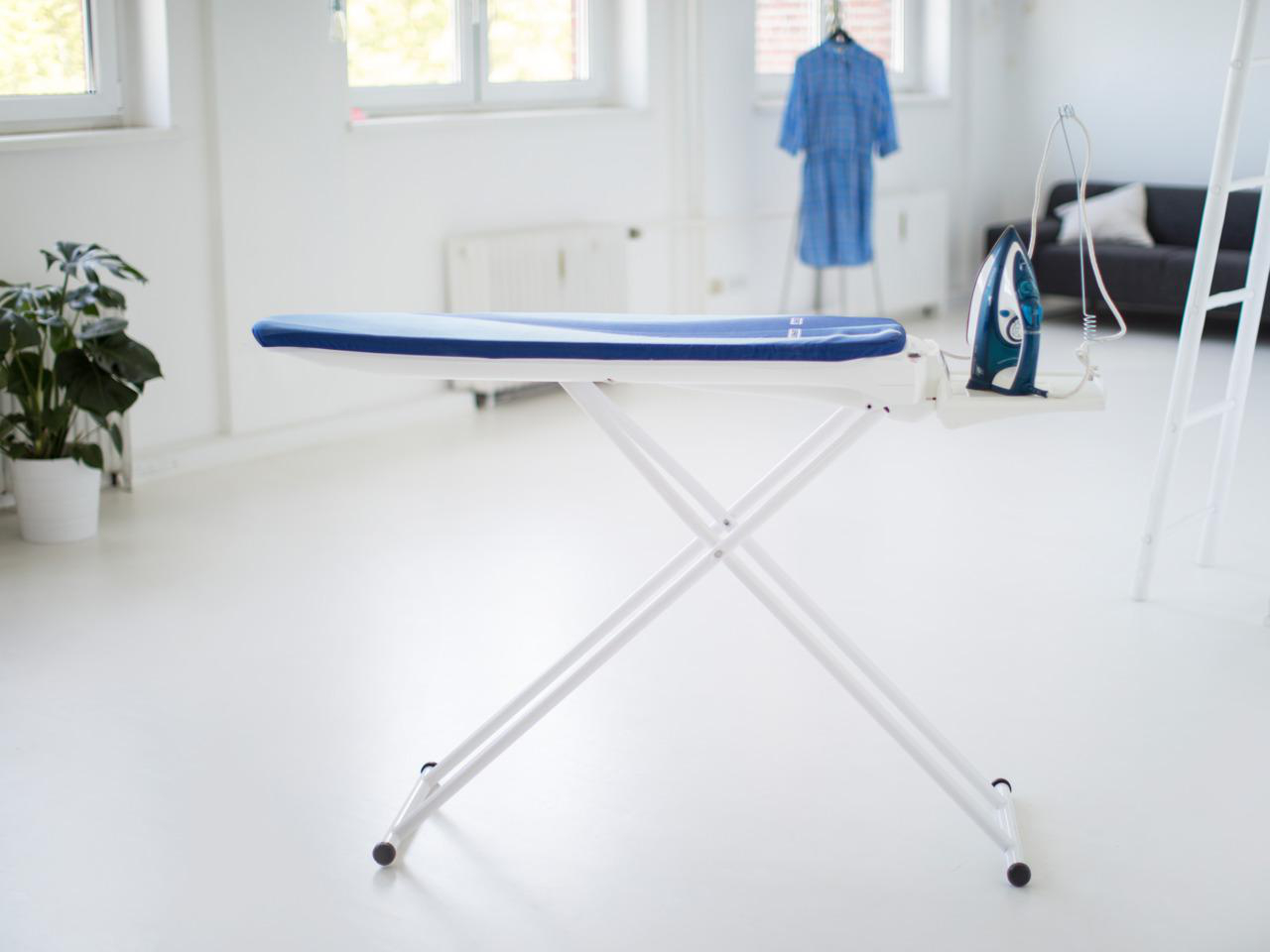 Bügelbrett mit blauem Bezug, Bügeleisen und Bügeleisenablage in hellem Raum.