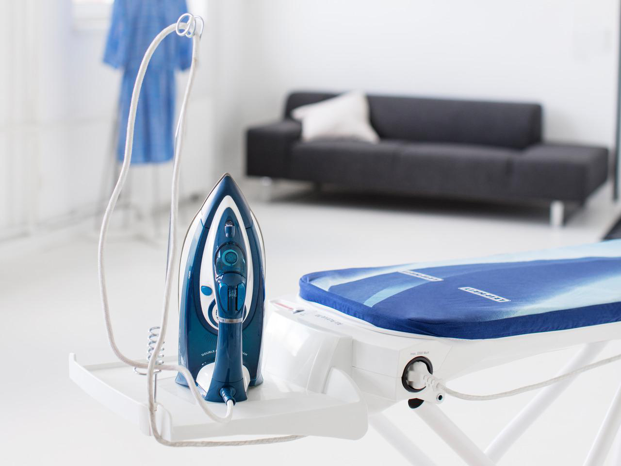 Ein Bügeleisen und ein Bügelbrett in einem weißen Raum. Ein Sofa und ein Kleidungsstück auf einem Ständer sind im Hintergrund.