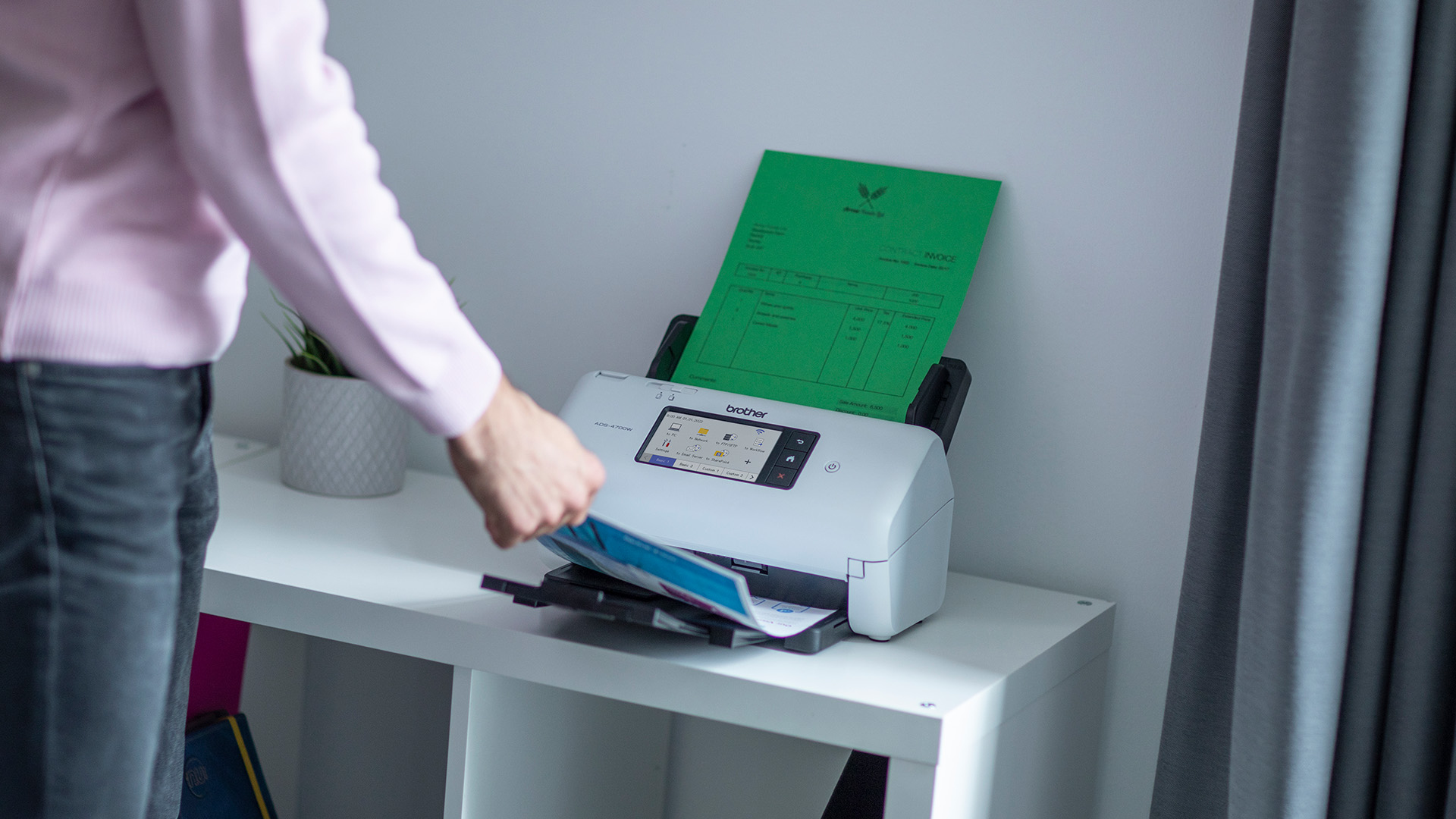 Una persona inserta un documento verde en un escáner Brother blanco sobre una mesa blanca. Una maceta cerca.