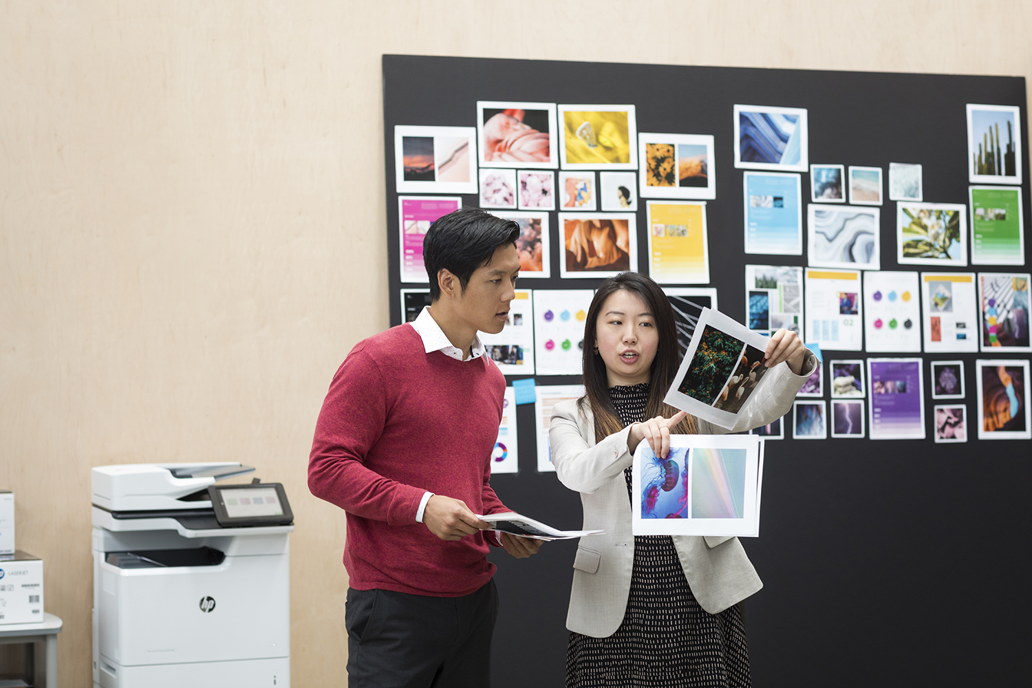 Deux personnes dans un bureau regardant des photos sur un mur. Une imprimante est visible.