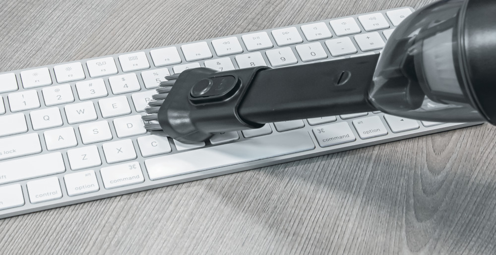 Un aspirateur noir nettoie un clavier d'ordinateur blanc sur une surface en bois.