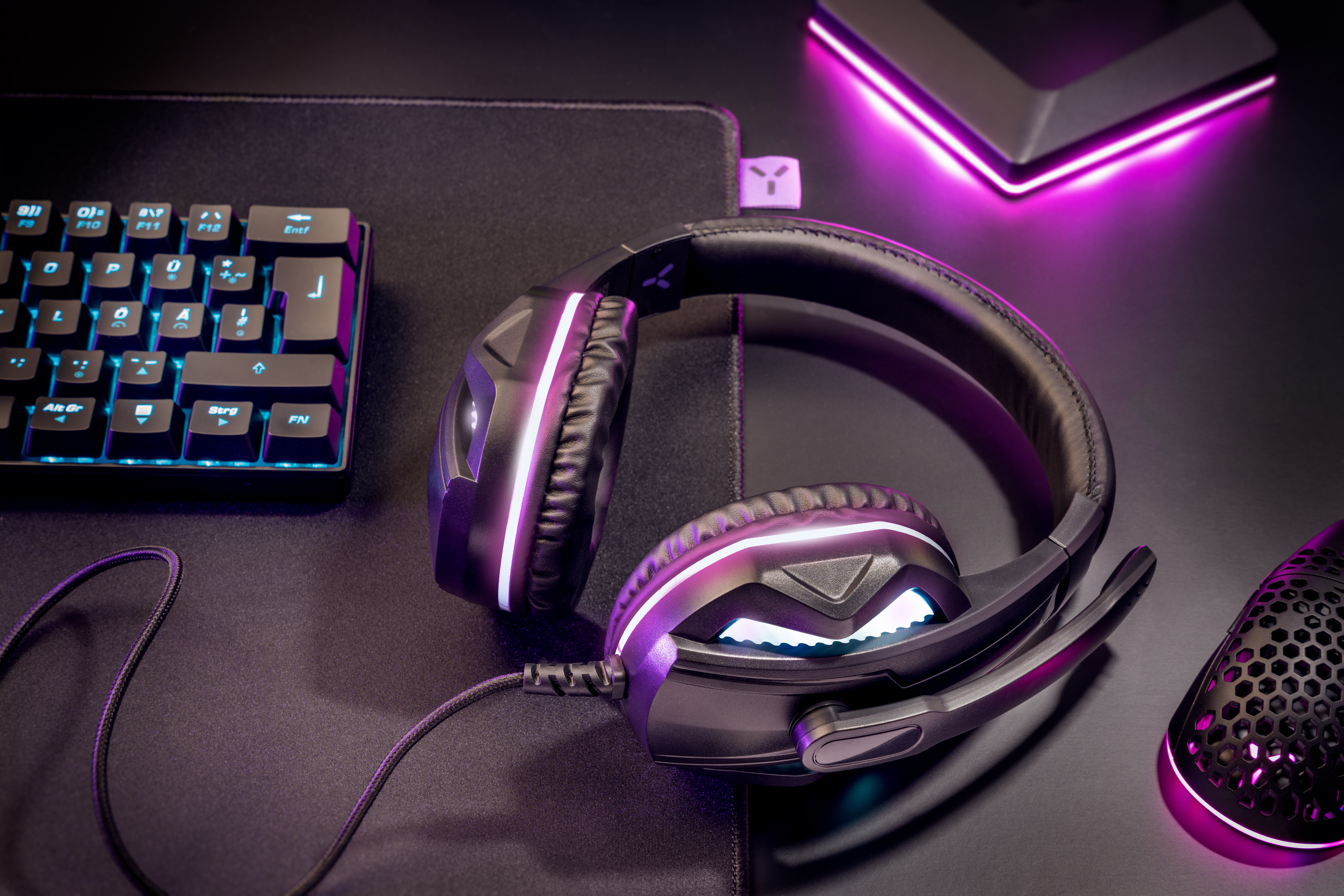 Gaming-Setup: Headset, Maus, Tastatur auf schwarzer Oberfläche mit lila Lichtern.
